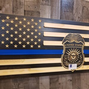 May include: Wooden American flag with a blue line and gold stars. The flag features a carved United States Federal Marshal badge. The flag is made of stained wood and has a rustic appearance.