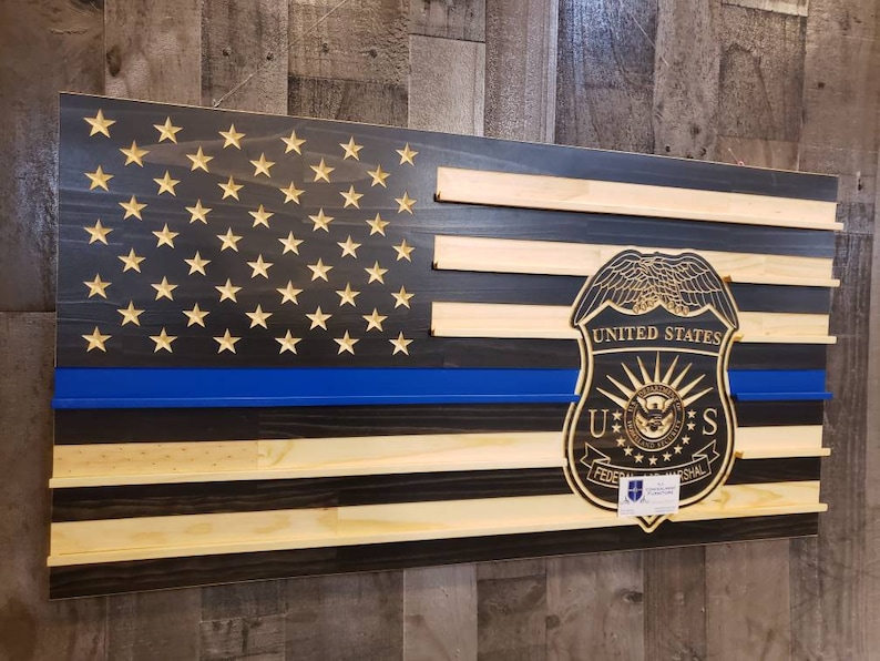 May include: Wooden American flag with a blue line and gold stars. The flag features a carved United States Federal Marshal badge. The flag is made of stained wood and has a rustic appearance.