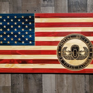 May include: A wooden American flag with a red, white, and blue color scheme. The flag has a black and gold circle in the lower right corner with the text "Initial Success or Total Failure" and a military insignia.