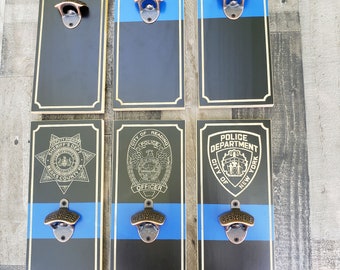 Puede incluir: Seis abridores de botellas de madera con una franja azul y varios logotipos de aplicación de la ley. Los logotipos incluyen el Departamento de Justicia, Saint Michael Protect Us, la Patrulla Estatal de Colorado, el Departamento del Sheriff del Condado de Berks, el Oficial de Policía de la Ciudad de Reading y el Departamento de Policía de la Ciudad de Nueva York.