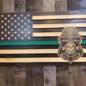 May include: Wooden American flag with gold stars, a green stripe, and a CBP Border Patrol badge. The flag is made of stained wood and has a rustic appearance. The badge is a detailed emblem.