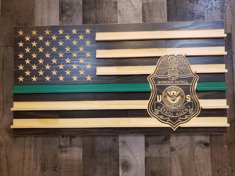 May include: Wooden American flag with gold stars, a green stripe, and a CBP Border Patrol badge. The flag is made of stained wood and has a rustic appearance. The badge is a detailed emblem.