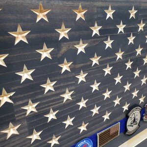 Op de afbeelding: Een Amerikaanse houten vlag met een verweerde donkerblauwe achtergrond en gouden sterren. De vlag heeft een blauw lint met een patch erop. Het lint is met een spijker aan de vlag bevestigd. De vlag hangt aan een muur.