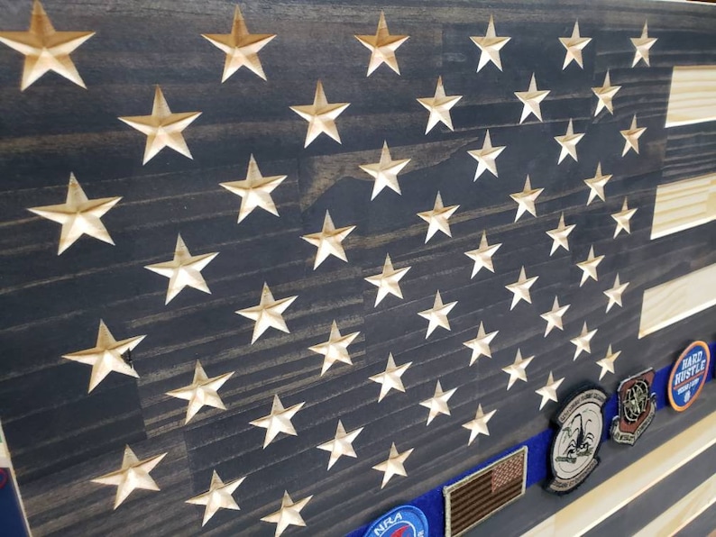 Op de afbeelding: Een Amerikaanse houten vlag met een verweerde donkerblauwe achtergrond en gouden sterren. De vlag heeft een blauw lint met een patch erop. Het lint is met een spijker aan de vlag bevestigd. De vlag hangt aan een muur.