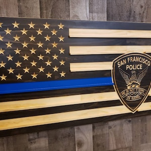 May include: A wooden American flag with a blue line, a thin blue line, and a San Francisco Police Department badge. The badge is a shield with an eagle and the words "San Francisco Police" and "Oro en Paz, Fierro en Guerra".