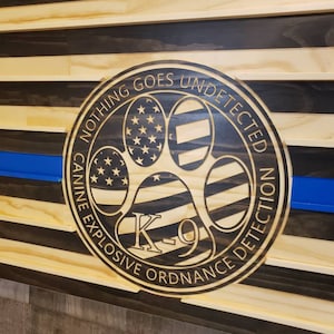 May include: A wooden American flag with a blue stripe and a round emblem in the lower right corner. The emblem features a paw print with the text "Nothing Goes Undetected Canine Explosive Ordnance Detection K-9".