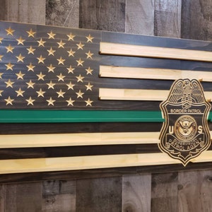 May include: Wooden American flag with carved stars and a green stripe. A CBP Border Patrol badge is affixed to the flag. The flag is made of wooden planks and has a rustic appearance.