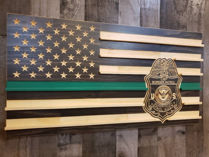 May include: Wooden American flag with carved stars and a green stripe. A CBP Border Patrol badge is affixed to the flag. The flag is made of wooden planks and has a rustic appearance.