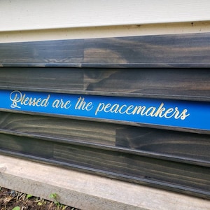 May include: A wooden wall shelf with a blue stripe and the text "Blessed are the peacemakers" in gold lettering. The shelf has five shelves for displaying items.