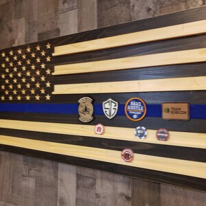 Op de afbeelding: Een Amerikaanse houten vlag met een blauwe streep in het midden. De vlag heeft een zwarte achtergrond en is versierd met verschillende patches, waaronder "Team Rubicon", "Hard Hustle" en een schild met een "G" erop.