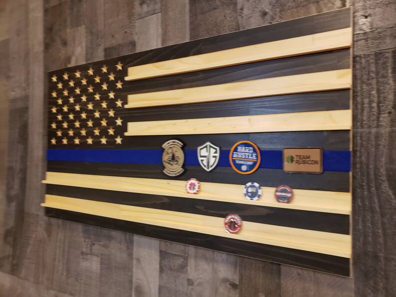 Op de afbeelding: Een Amerikaanse houten vlag met een blauwe streep in het midden. De vlag heeft een zwarte achtergrond en is versierd met verschillende patches, waaronder "Team Rubicon", "Hard Hustle" en een schild met een "G" erop.