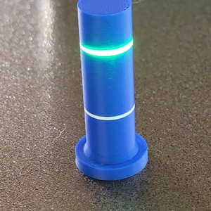 May include: A blue cylindrical object with a circular base and a flat top. The object has two white bands and a glowing green band. The object is on a textured surface.