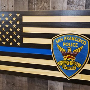 May include: A wooden American flag with a blue line and the San Francisco Police Department logo. The logo is blue with a gold eagle and the words "San Francisco Police" in gold. The words "Oro en Paz, Fierro en Guerra" are written in gold on a banner below the eagle.