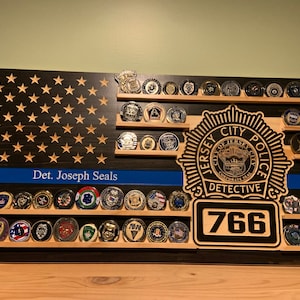 K&ouml;nnte beinhalten: Holz-Display in Form einer amerikanischen Flagge mit Regalen, die verschiedene Sammlerm&uuml;nzen halten. Das Flaggen-Design zeigt eine blaue Linie, Sterne und den Text "Det. Joseph Seals". Ein Abzeichen des Jersey City Police Detective und die Nummer 766 sind ebenfalls zu sehen.