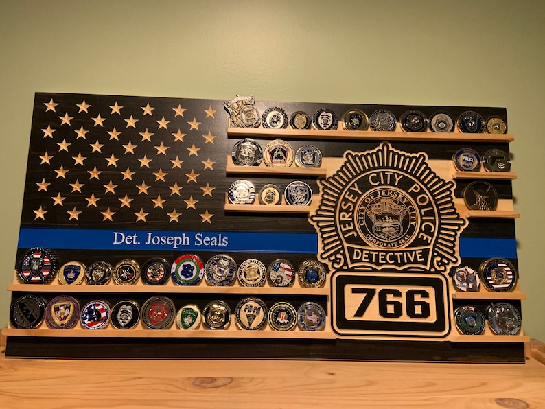 K&ouml;nnte beinhalten: Holz-Display in Form einer amerikanischen Flagge mit Regalen, die verschiedene Sammlerm&uuml;nzen halten. Das Flaggen-Design zeigt eine blaue Linie, Sterne und den Text "Det. Joseph Seals". Ein Abzeichen des Jersey City Police Detective und die Nummer 766 sind ebenfalls zu sehen.