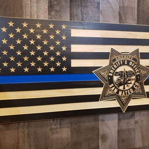 K&ouml;nnte beinhalten: Amerikanische Holzflagge mit blauer Linie und Sheriff-Abzeichen. Die Flagge zeigt geschnitzte Sterne und Streifen, mit dem Emblem des Sheriff-B&uuml;ros von San Mateo County.