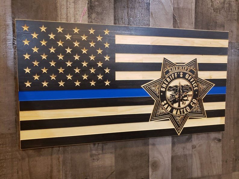 K&ouml;nnte beinhalten: Amerikanische Holzflagge mit blauer Linie und Sheriff-Abzeichen. Die Flagge zeigt geschnitzte Sterne und Streifen, mit dem Emblem des Sheriff-B&uuml;ros von San Mateo County.
