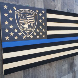 Op de afbeelding: Een Amerikaanse vlag van hout met een blauwe lijn, die de rechtshandhaving vertegenwoordigt, en een aangepast schild met de tekst "East Providence Police K9 Patrol-Narcotic RI".