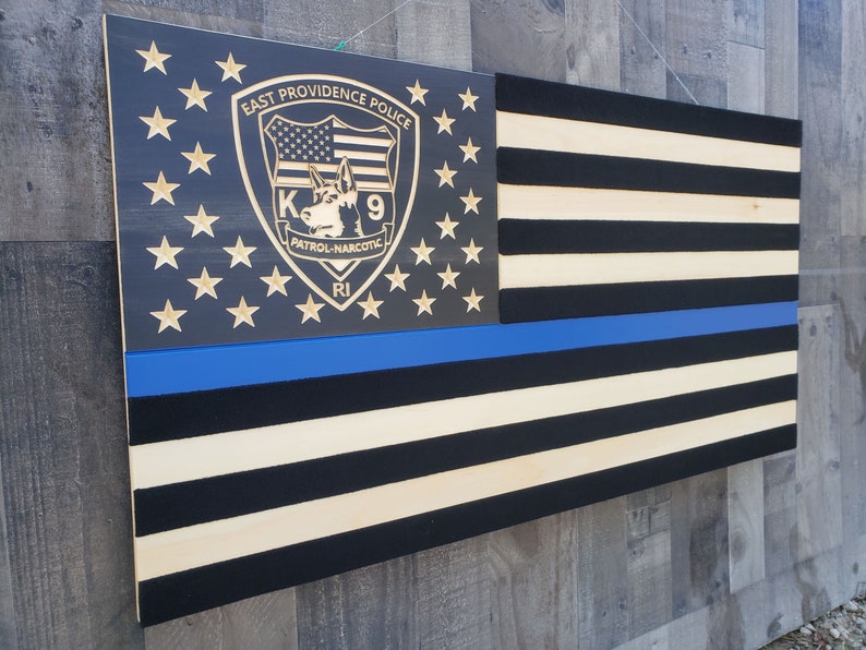 Op de afbeelding: Een Amerikaanse vlag van hout met een blauwe lijn, die de rechtshandhaving vertegenwoordigt, en een aangepast schild met de tekst "East Providence Police K9 Patrol-Narcotic RI".