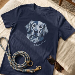 May include: A navy blue short-sleeved t-shirt featuring a stylized graphic of a golden retriever's face in shades of blue and white, with the text "Goldie Mom" printed below the dog's chin, accompanied by a coiled blue and tan rope leash.