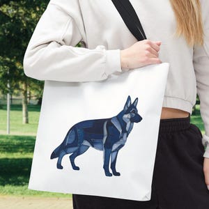 Puede incluir: Bolso tote blanco con una ilustración estilizada de un perro pastor alemán en mezclilla azul. El perro está representado en un estilo geométrico de patchwork. El bolso tiene una correa negra.