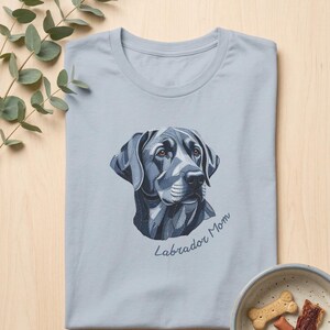 May include: A light blue t-shirt featuring a stylized graphic of a Labrador retriever head in shades of blue and grey, with the text "Labrador Mom" written in a cursive script underneath the dog illustration on the front of the garment.