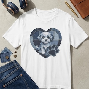 Op de afbeelding: Een wit T-shirt met korte mouwen en een centrale afbeelding van een puppy die in een hartvormig denim patchworkontwerp zit, vergezeld van een donkerblauwe pootafdruk. Het shirt wordt getoond naast een spijkerbroek, koptelefoon en een notitieboek.