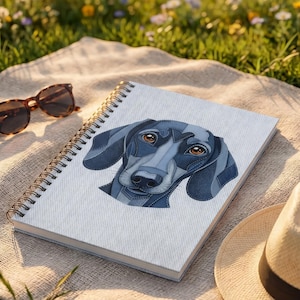 May include: A spiral-bound notebook featuring a stylized illustration of a dachshund dog's face in shades of blue and gray. The notebook is resting on a beige blanket, with sunglasses and a straw hat nearby. The scene is set outdoors with a blurred background of flowers and grass.
