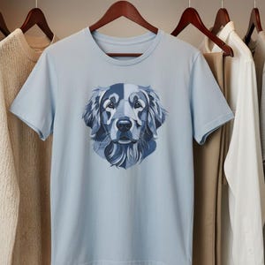 Op de afbeelding: Een lichtblauw t-shirt met korte mouwen en een grafische print van een hondenkop in blauwtinten. De hondenillustratie is opgebouwd uit geometrische vormen en lijnen, wat een gestileerd portret vormt van een hond met lang haar en flaporen.