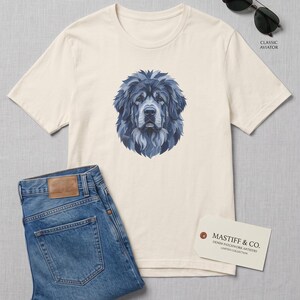 May include: A cream-coloured short-sleeve t-shirt featuring a blue graphic illustration of a dog's head, paired with blue denim jeans, black aviator sunglasses, and a white tag labelled MASTIFF & CO. DENIM PATCHWORK ARTISTRY LIMITED COLLECTION.