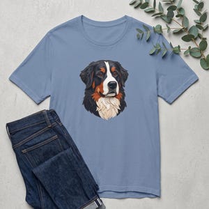May include: A dusty blue short-sleeved t-shirt featuring a centered graphic illustration of a Bernese Mountain Dog head with black, white, and tan fur, paired with a pair of dark blue denim jeans.
