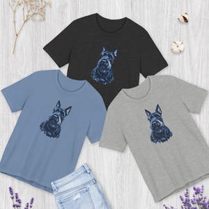 Puede incluir: Tres camisetas de manga corta y cuello redondo en gris carbón, azul polvoriento y gris jaspeado, cada una con una ilustración gráfica centrada de un perro Terrier escocés negro. Unos vaqueros azul claro se sitúan debajo de las camisetas.