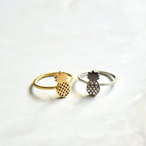 Puede incluir: Dos anillos de piña, uno dorado y otro plateado, sobre una superficie de mármol blanco.