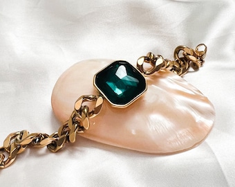 May include: A gold chain bracelet with a large, square, emerald green gemstone. The bracelet is on a white, silky surface.
