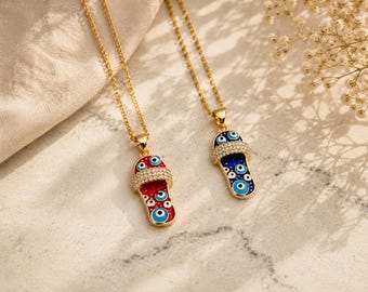 May include: Two gold-colored necklaces with pendant charms. Each charm is a sandal shape with a gold border, adorned with blue eye designs. One sandal is red, the other is blue, both with a crystal-studded strap.
