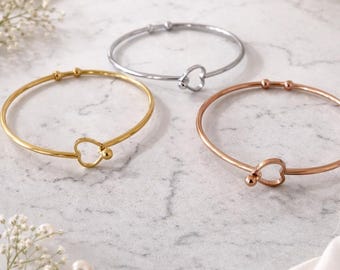 May include: Three delicate bracelets in gold, silver, and rose gold, each featuring a heart-shaped charm. The bracelets are displayed on a marble surface with white fabric and floral accents, creating an elegant presentation.