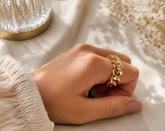 May include: A gold-toned ring featuring a thick, interlocking curb chain link design. The ring is worn on a finger, showcasing the polished, reflective metal surface and the repeating oval-shaped links that form the continuous band structure.