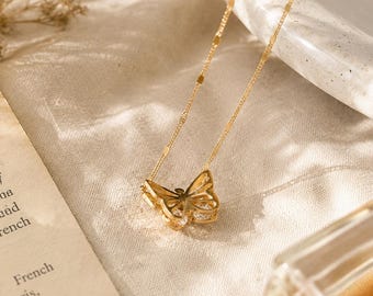 May include: A delicate gold-colored butterfly pendant necklace. The butterfly has intricate cutouts and is adorned with small, sparkling stones. The necklace chain is thin and elegant, with a subtle design. The necklace rests on a soft, textured surface.