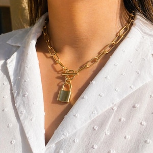May include: Gold-toned chain necklace with a padlock pendant. The necklace features a chunky link chain and a toggle clasp. The padlock pendant hangs from a small loop and has a rectangular shape. The necklace is worn over a white, textured blouse.