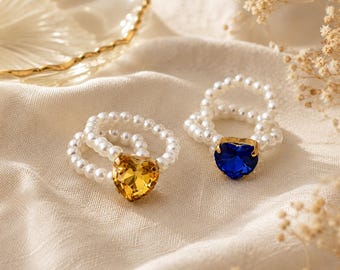 May include: Two rings featuring bands made of small white spherical beads. One ring has a faceted heart-shaped yellow gemstone set in gold-tone metal, and the other ring has a faceted heart-shaped blue gemstone set in gold-tone metal.