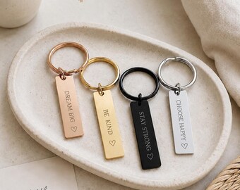 May include: Four rectangular metal keychains with circular rings. The rose gold keychain reads DREAM BIG, the gold keychain reads BE KIND, the black keychain reads STAY STRONG, and the silver keychain reads CHOOSE HAPPY, each with a heart symbol.
