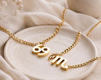 May include: Two gold-toned metal necklaces featuring pendants on a curb chain. One pendant displays the number 69, and the other pendant displays the lowercase letter m. The necklaces are crafted from a polished, reflective gold-colored metal material.