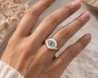 May include: A gold-toned signet ring featuring a white oval inlay with a central turquoise stone surrounded by small clear crystals, forming an eye motif. The ring has a polished metallic band and is worn on a finger, showcasing the detailed design.
