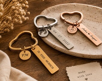 May include: Three heart-shaped keychains in gold, silver, and rose gold tones. Each features a rectangular tag and a small circular charm. Engravings include dates, names, initials, footprints, and the text "made with love" on a separate paper card.