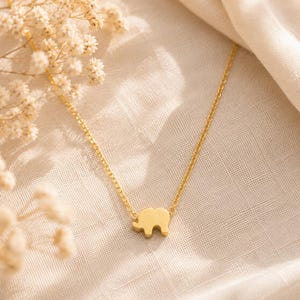 May include: A gold-toned necklace featuring a small, flat elephant-shaped pendant suspended from a delicate, thin link chain. The pendant is centered on the chain, which has a polished, reflective surface that catches the light.