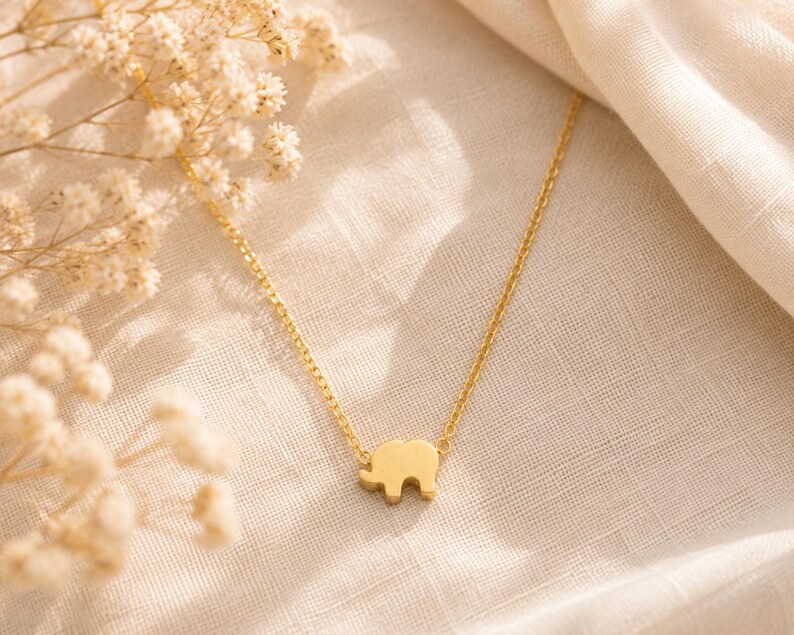 May include: A gold-toned necklace featuring a small, flat elephant-shaped pendant suspended from a delicate, thin link chain. The pendant is centered on the chain, which has a polished, reflective surface that catches the light.