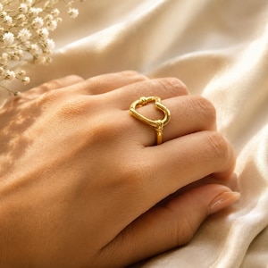 May include: A gold-colored ring with an abstract heart-shaped design. The ring is worn on a hand, with a soft, neutral-toned background. The ring's design features a looped shape with a knot detail, showcasing a modern jewelry piece.