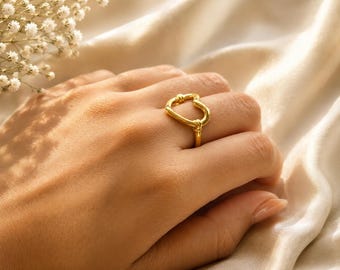 May include: A gold-colored ring with an abstract heart-shaped design. The ring is worn on a hand, with a soft, neutral-toned background. The ring's design features a looped shape with a knot detail, showcasing a modern jewelry piece.