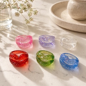 May include: Six colorful resin rings with glitter. The rings are in shades of pink, purple, clear, red, green, and blue. Each ring has a heart-shaped design. The rings are displayed on a marble surface.