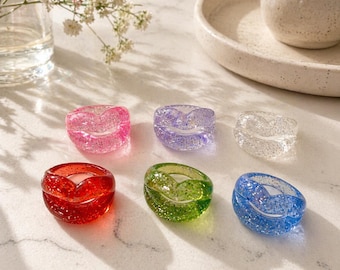 May include: Six colorful resin rings with glitter. The rings are in shades of pink, purple, clear, red, green, and blue. Each ring has a heart-shaped design. The rings are displayed on a marble surface.